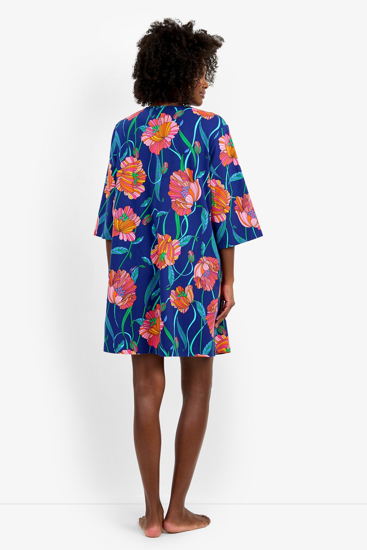 A person stands barefoot facing away, wearing the Linear Poppy Sleepshirt—a blue dress featuring large, colorful orange and pink floral patterns and wide sleeves. The background is plain white.