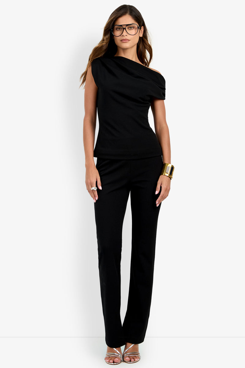 A woman stands against a plain white background wearing the Roza Top, an asymmetrical black short-sleeve top, paired with black pants, a gold bracelet, glasses, and strappy silver heels.