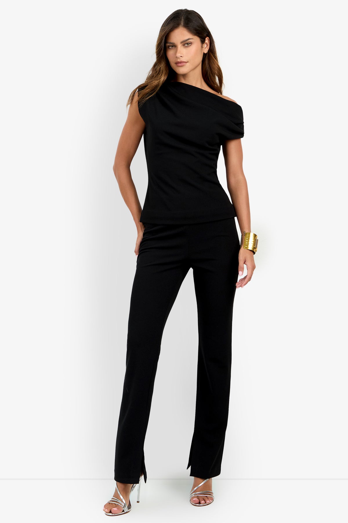 Woman stands wearing the Roza Top, a stylish black asymmetrical piece, paired with black pants featuring small front slits, a gold cuff bracelet, and silver high-heeled sandals, posing against a plain white background.