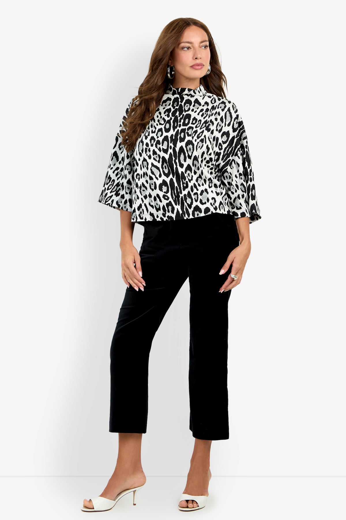 A woman wearing the Sidony Top—a black and white leopard print blouse with wide sleeves—paired with black cropped pants, white heeled sandals, and large earrings, stands against a plain white background.