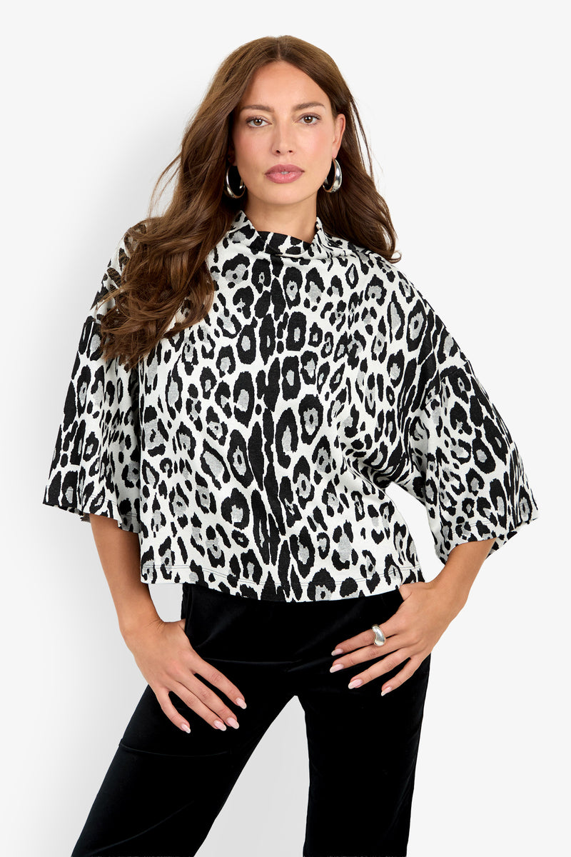 A woman with long brown hair stands against a white background, wearing the Sidony Top—a black-and-white leopard print style with wide sleeves—paired with black pants and posing with one hand on her hip.