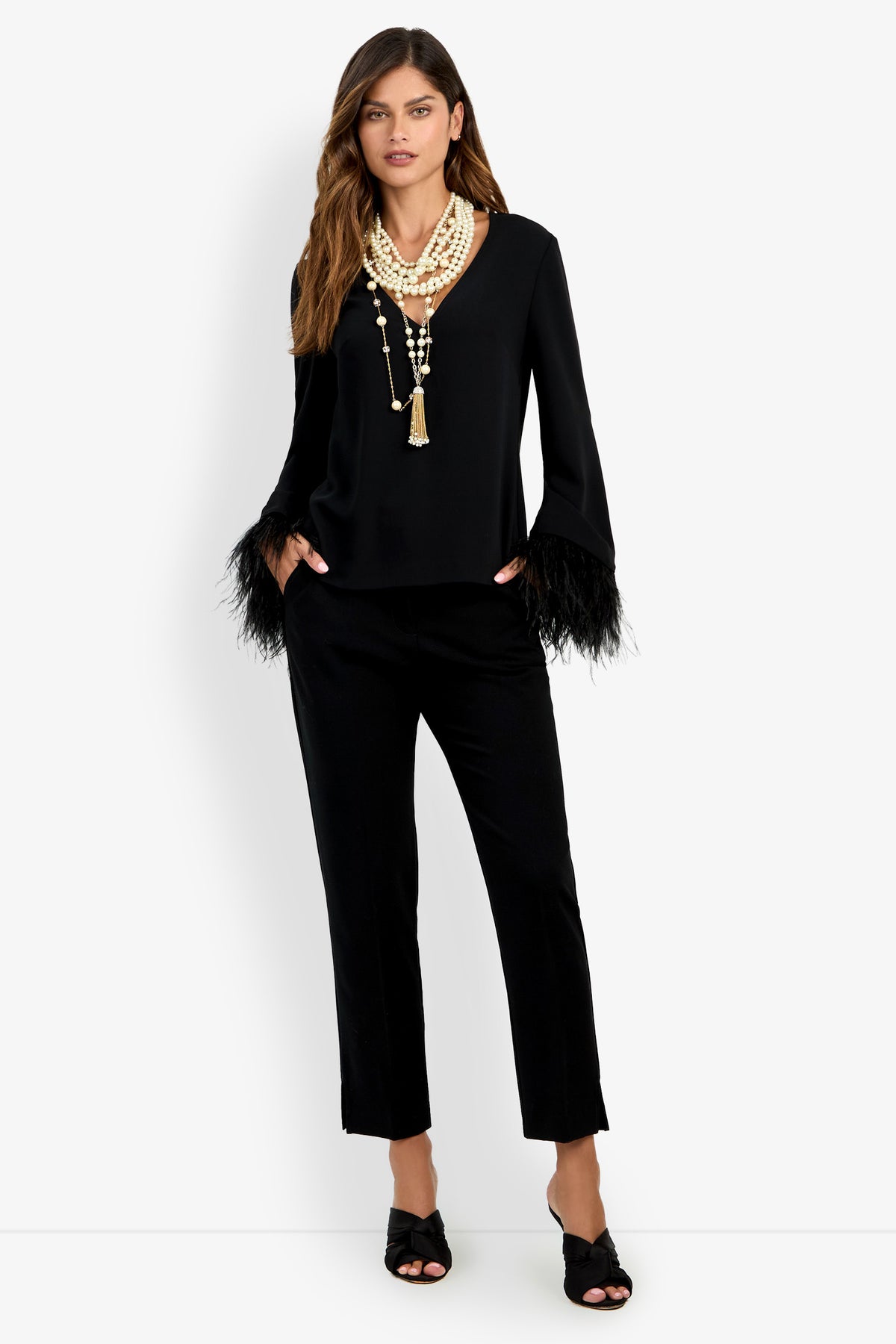 A woman wears the Anina Top, a black blouse featuring feathered sleeve cuffs, paired with black cropped pants, black open-toe heels, and layered pearl necklaces. She stands with her hands in her pockets against a plain white background.
