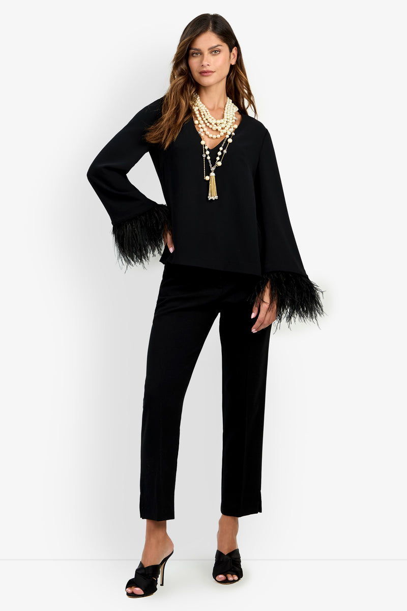 A woman stands confidently against a plain white background, wearing the Anina Top—a black outfit with feathered sleeves—paired with black heeled sandals and layered pearl necklaces. She has long brown hair.