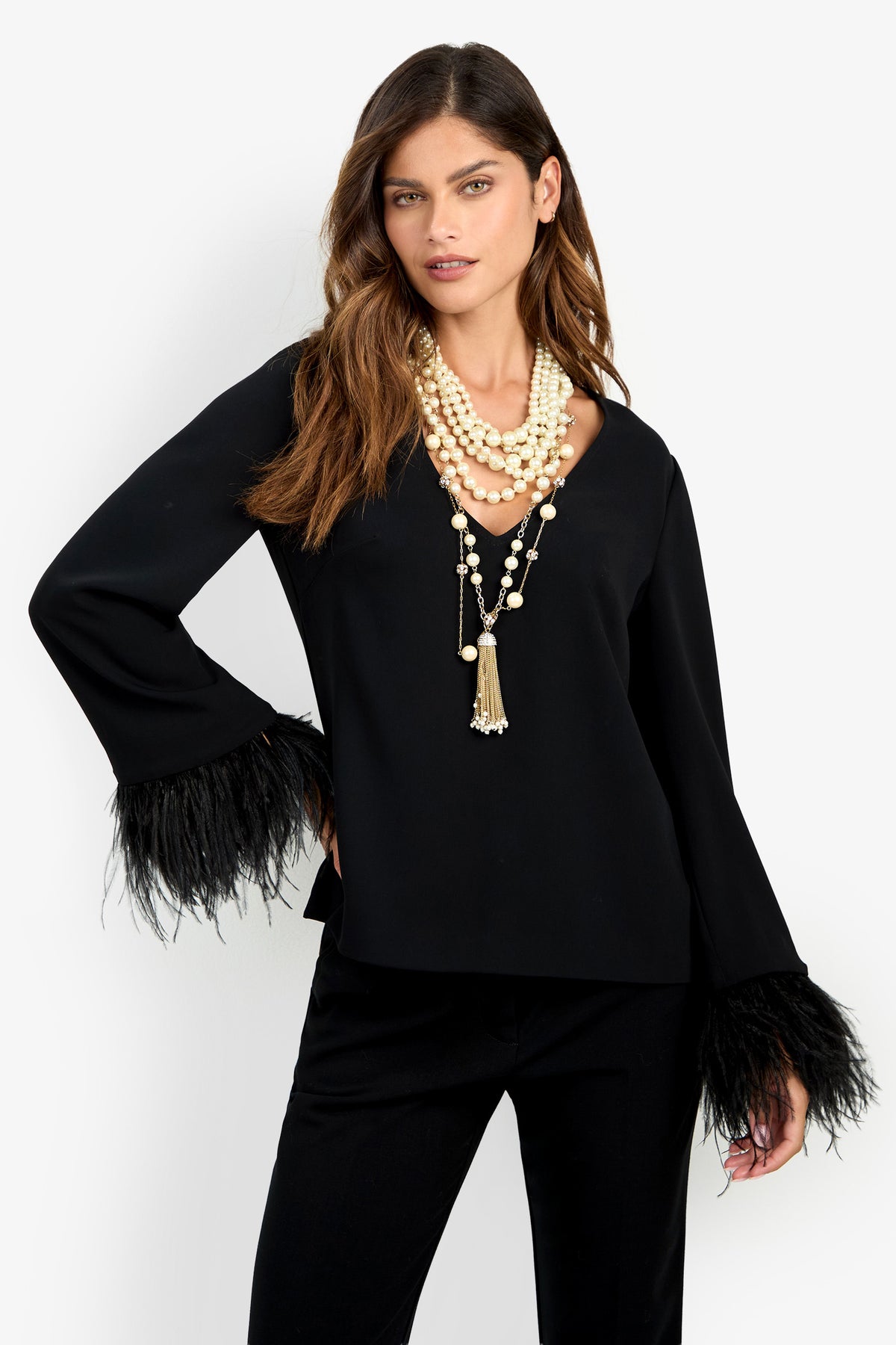 A woman wearing the Anina Top with feathered cuffs, paired with black pants and layered pearl necklaces, stands against a plain white background.