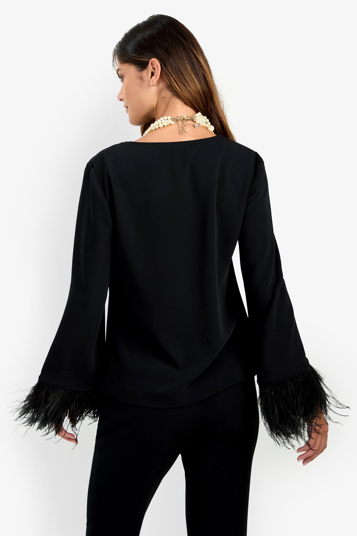 A woman faces away, wearing the Anina Top—a black long-sleeve shirt with feathered cuffs—paired with black pants and a chunky pearl necklace against a white background.