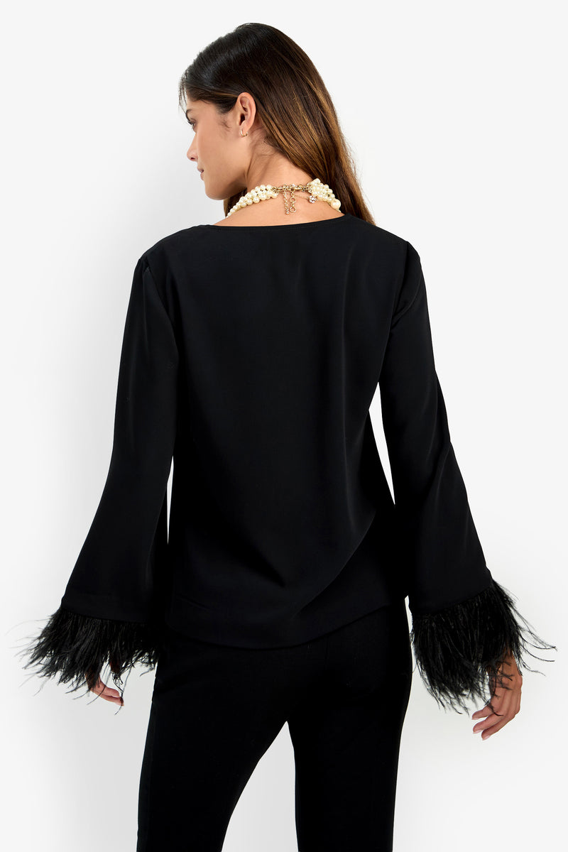 A woman faces away, wearing the Anina Top—a black long-sleeve shirt with feathered cuffs—paired with black pants and a chunky pearl necklace against a white background.