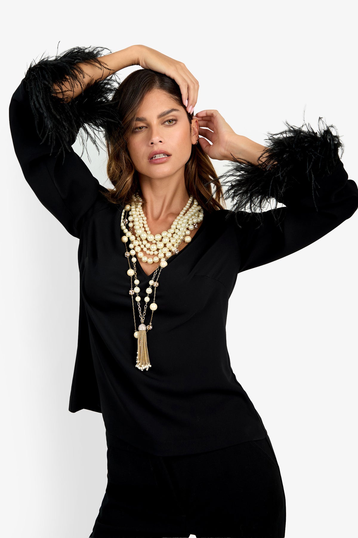 Woman posing in the Anina Top, a black long-sleeve design with feathered cuffs, styled with layered pearl necklaces and set against a plain white background.