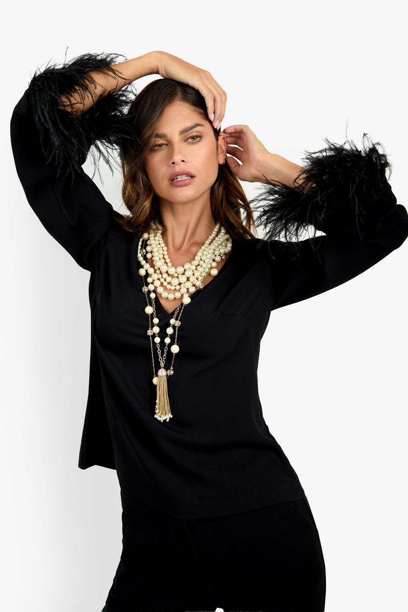 Woman posing in the Anina Top, a black long-sleeve design with feathered cuffs, styled with layered pearl necklaces and set against a plain white background.