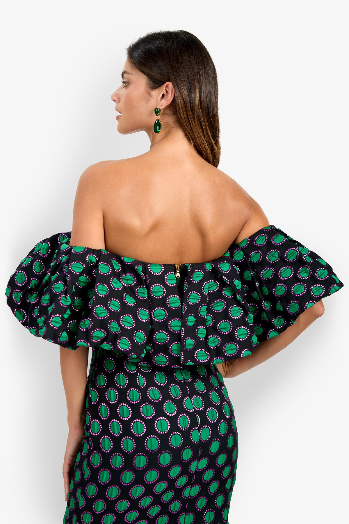 Wearing the Dafinas Top, a woman faces away, revealing her bare back and long brown hair. The off-shoulder, ruffled design features green and purple circular patterns, complemented by matching green drop earrings.