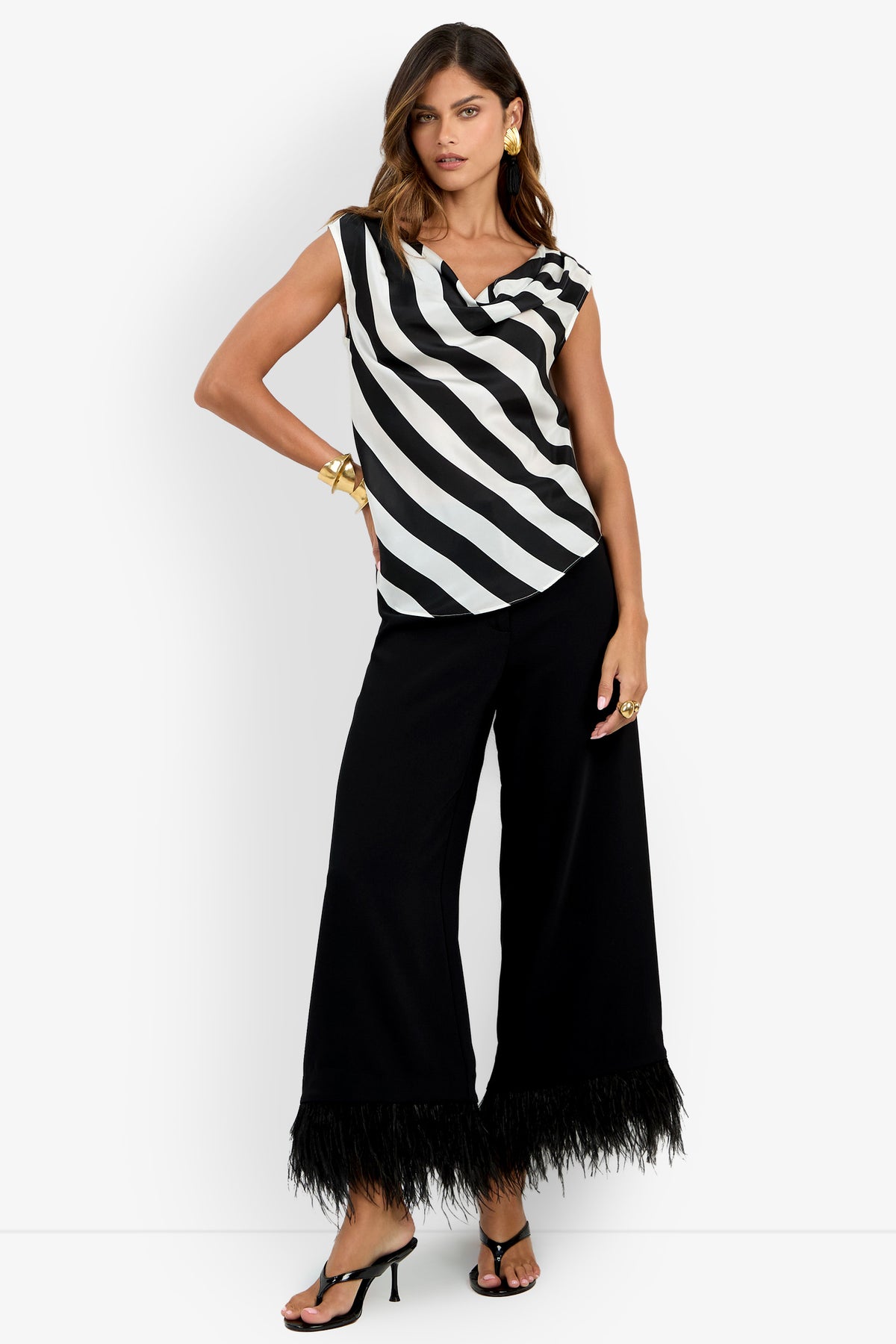 Woman wearing the Alessi Top—a black and white diagonal striped sleeveless top—paired with wide-leg black pants with feathered hems, black heeled sandals, and gold jewelry, standing against a white background.