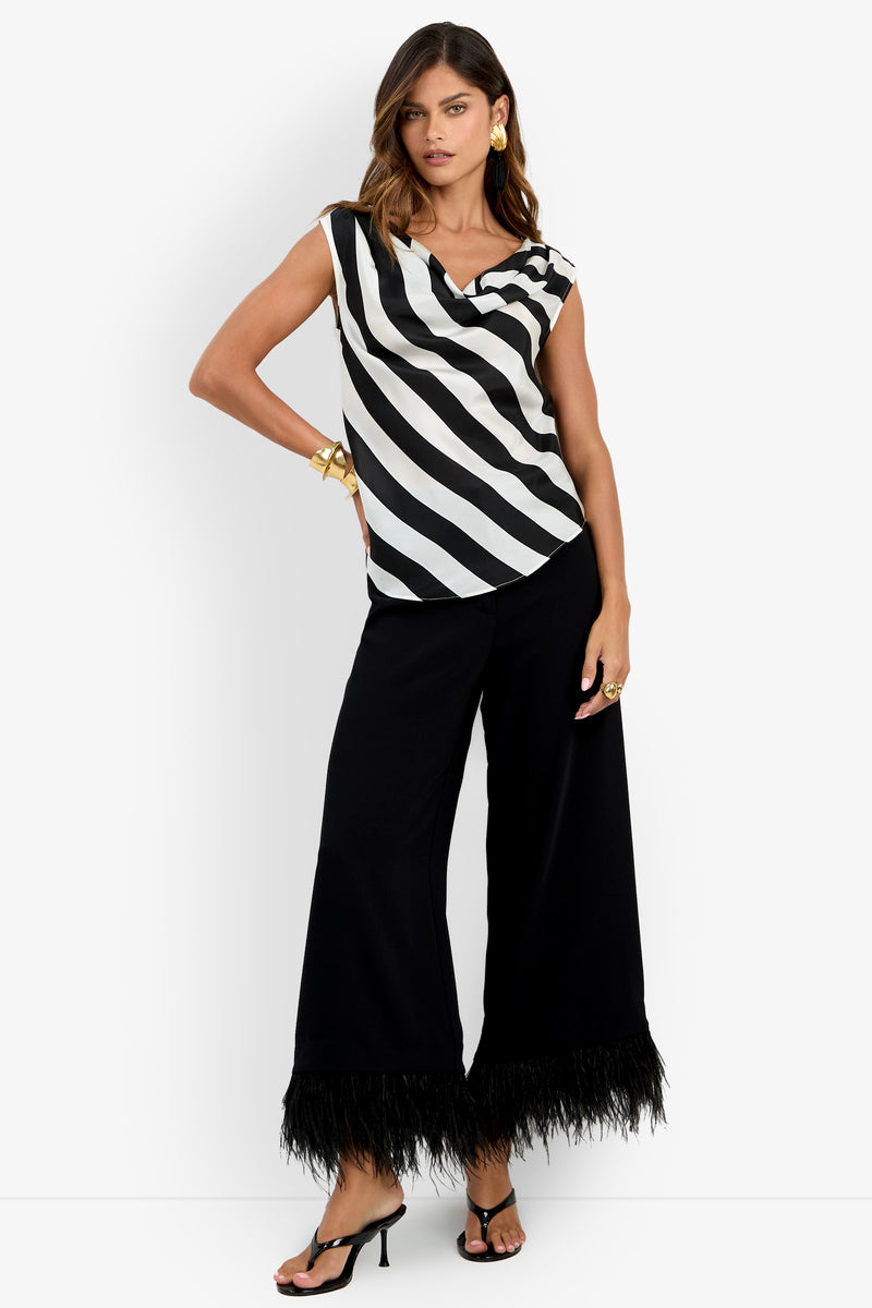 Woman wearing the Alessi Top—a black and white diagonal striped sleeveless top—paired with wide-leg black pants with feathered hems, black heeled sandals, and gold jewelry, standing against a white background.