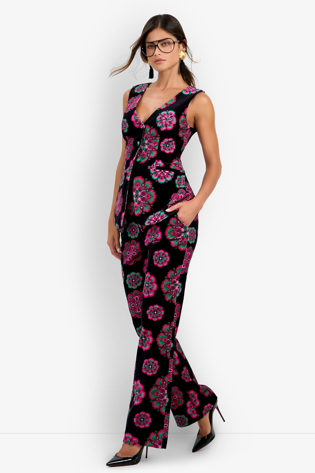 A woman wearing glasses and the Raylee Top—a sleeveless, fitted black top with pink and teal floral patterns—paired with black pointed heels and earrings, standing against a white background with her hands in her pockets.