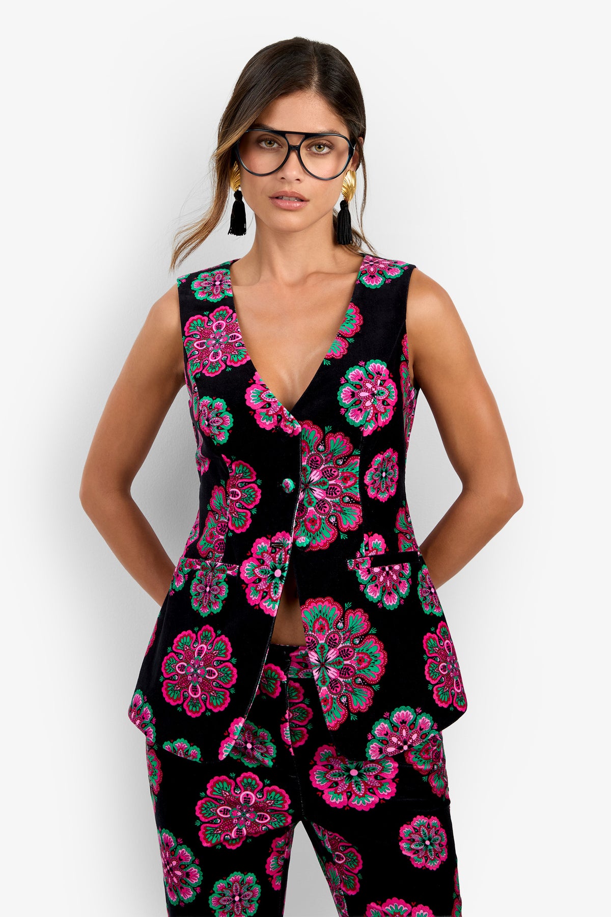 Woman wearing the Raylee Top, a black sleeveless suit adorned with bright pink, green, and blue floral patterns. She pairs it with matching pants, black glasses, and tassel earrings while standing against a plain white background with her hands behind her back.
