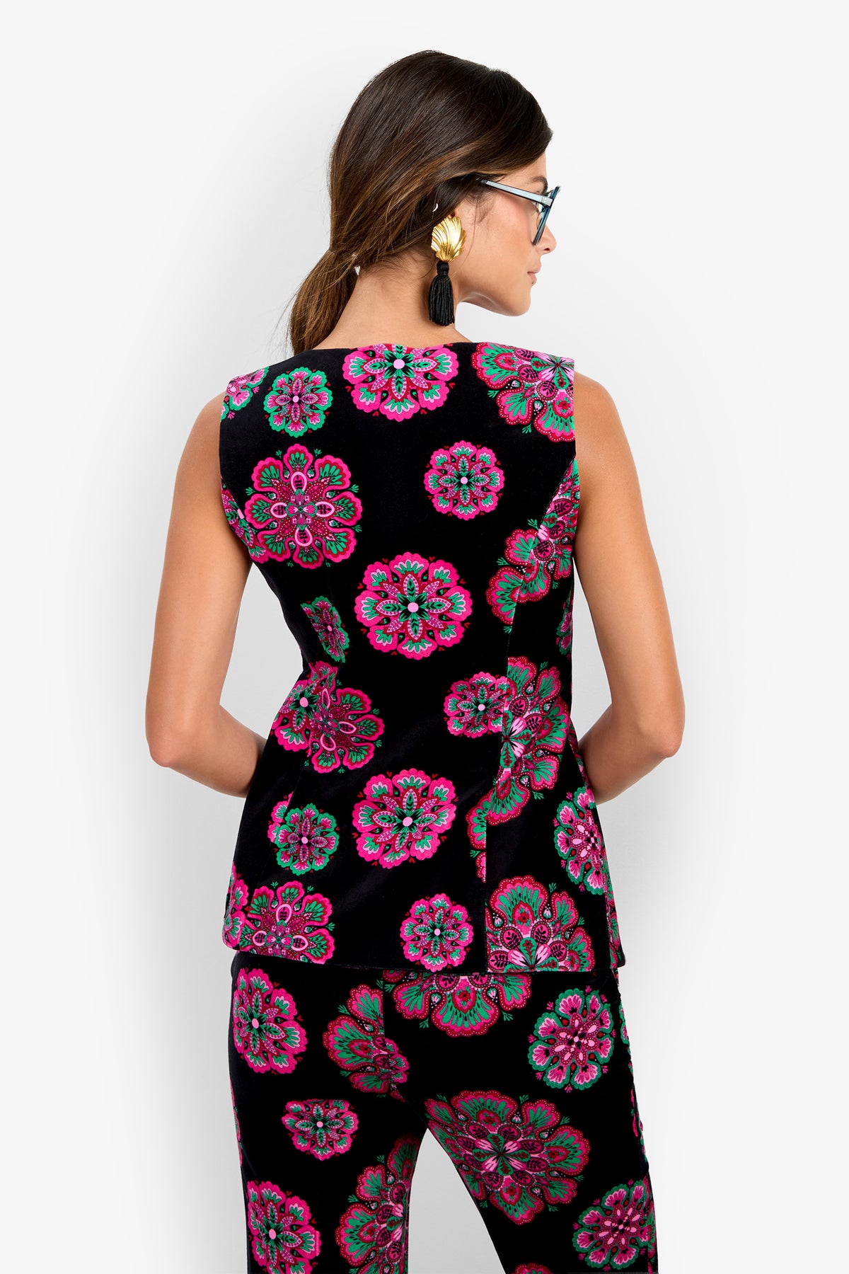 Woman facing away, wearing the sleeveless Raylee Top and matching pants with bright pink and green floral patterns, statement earrings, and glasses, standing against a plain white background.