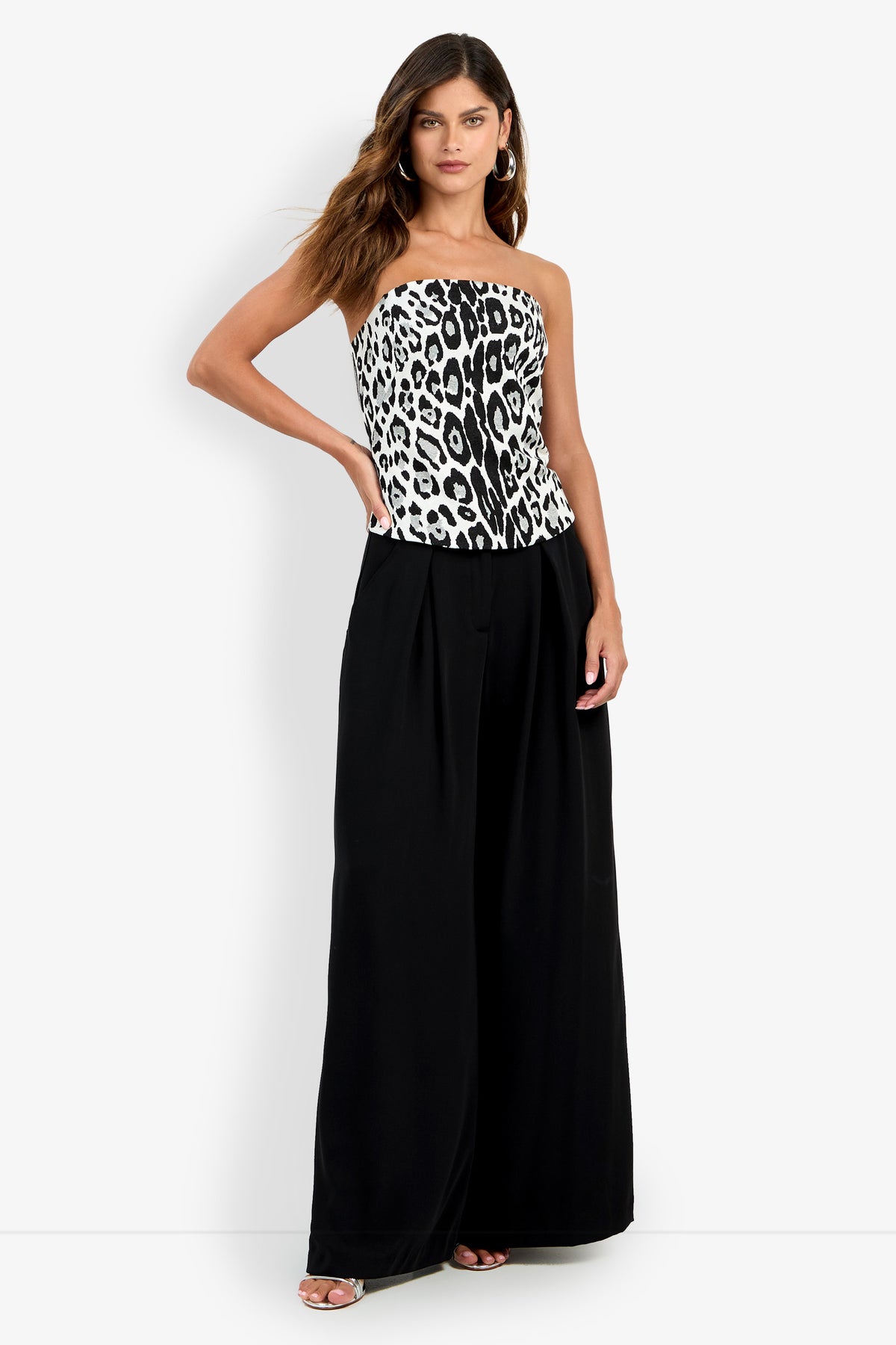 A woman stands confidently against a plain background, wearing the Mirisa Top in MULTI—a strapless, black and white leopard print style—paired with high-waisted, wide-leg black pants.