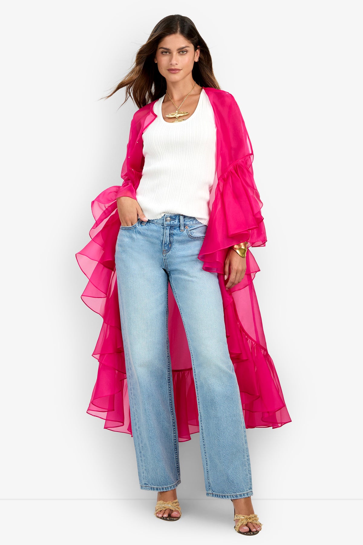 A woman stands wearing the Aries Jacket—a bright pink sheer ruffled duster—over a white top, blue jeans, and gold sandals. She has long brown hair and accessorizes with layered necklaces and a bracelet.