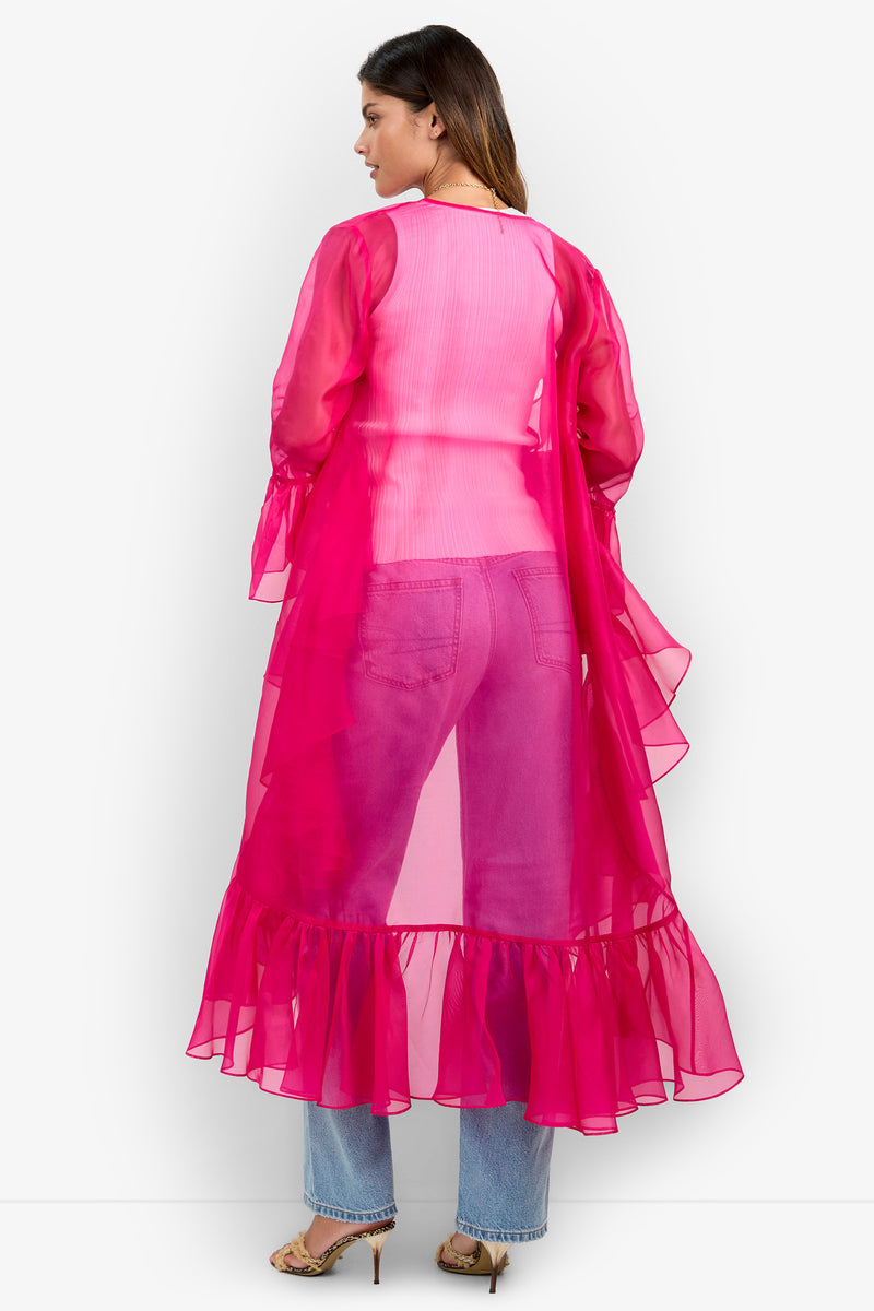 A woman stands facing away, wearing the Aries Jacket—a sheer hot pink ruffled long shrug—over a light pink top and blue jeans, paired with beige heels.