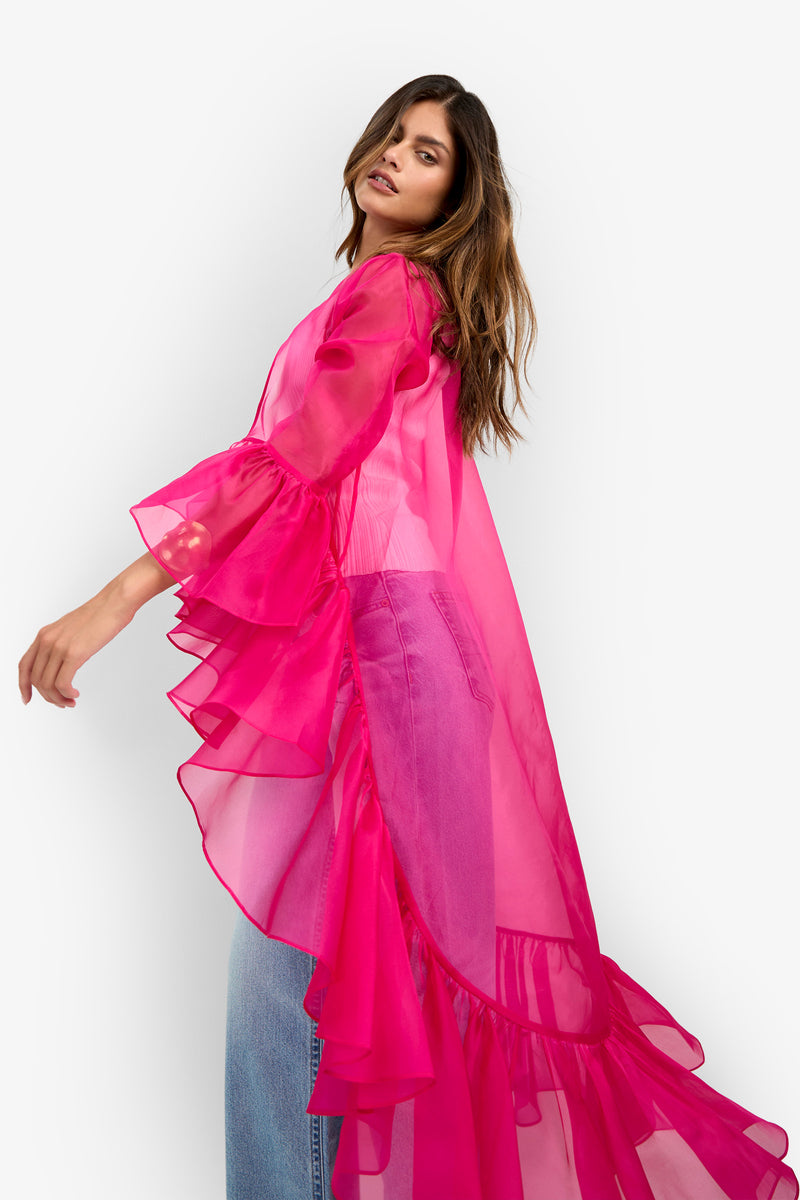 Wearing the Aries Jacket—a sheer, bright pink ruffled long top—over light blue jeans, a woman poses against a white background, looking over her shoulder.