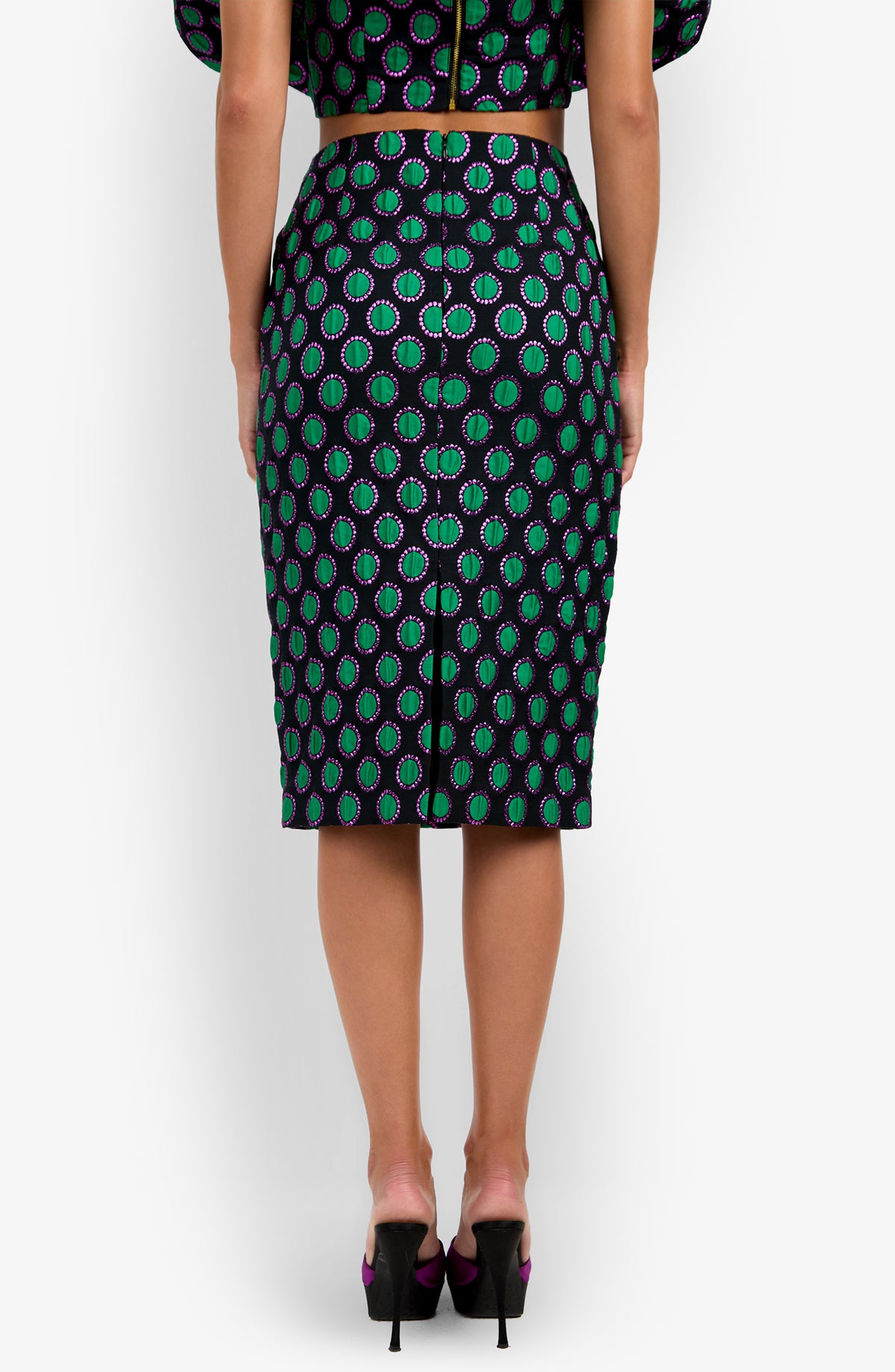 A woman stands facing away, wearing the fitted, knee-length Soniah Skirt with a green, purple, and black circular pattern. The skirt features a visible zipper down the back, and she pairs it with black high heels.