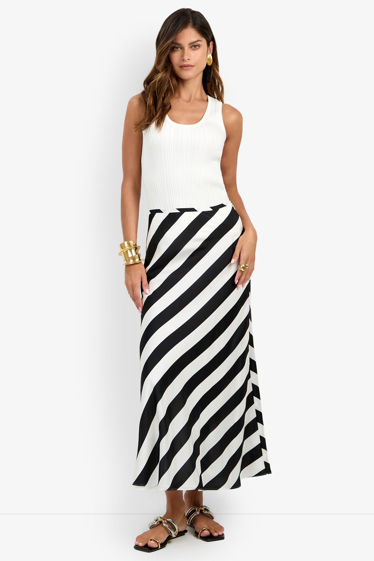 Wearing the Rabina Skirt—a long black-and-white diagonal striped design—a woman styled in a sleeveless white top, gold jewelry, and sandals stands against a plain white background.