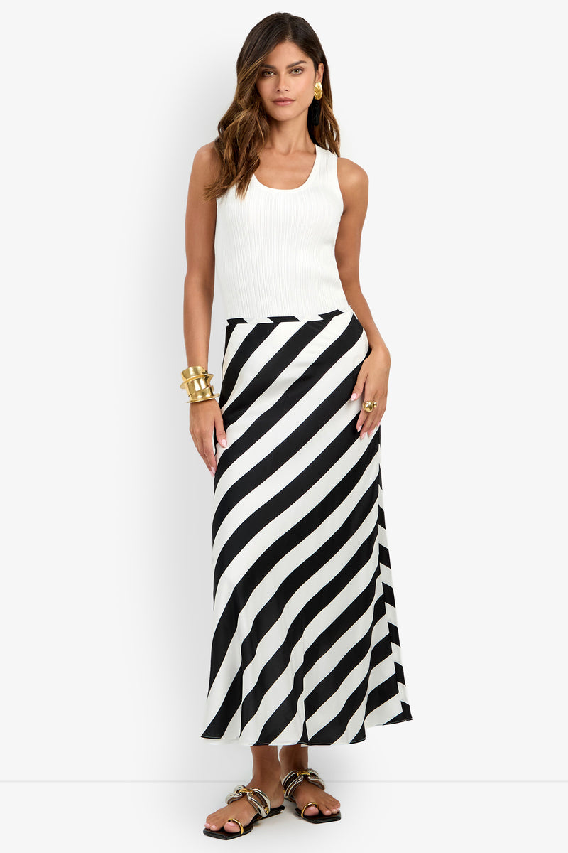 Wearing the Rabina Skirt—a long black-and-white diagonal striped design—a woman styled in a sleeveless white top, gold jewelry, and sandals stands against a plain white background.