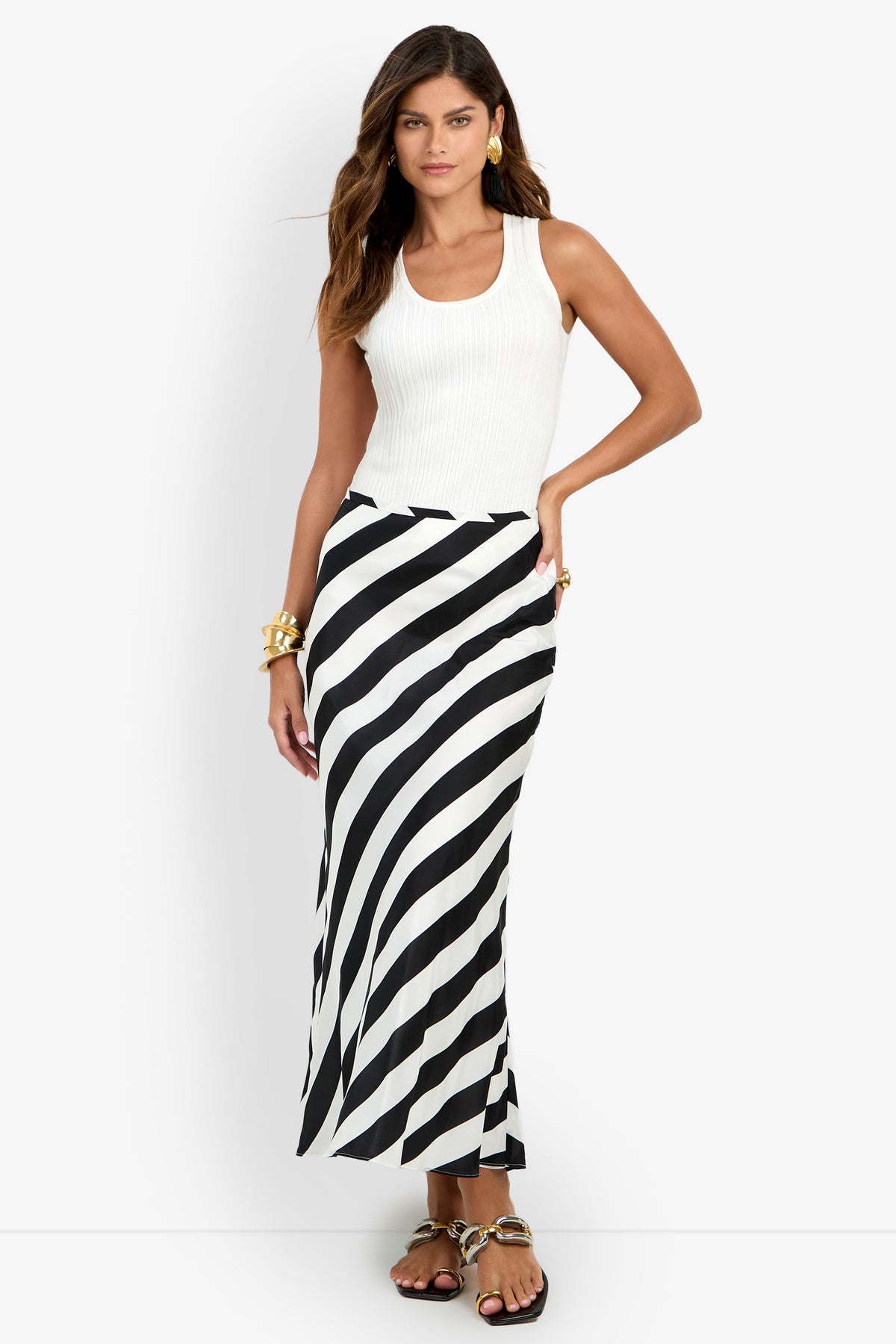 A woman models the Rabina Skirt—a long black-and-white diagonal striped skirt—paired with a sleeveless white top, gold jewelry, and sandals, standing against a plain white background.