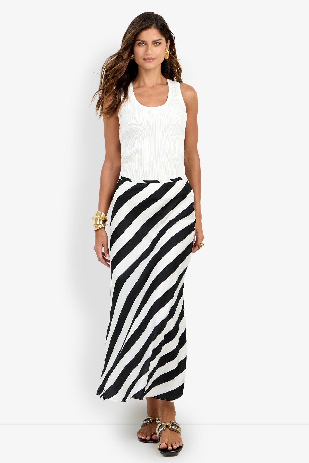 A woman stands against a plain white background wearing a white sleeveless top paired with the Rabina Skirt, a long black-and-white diagonal striped skirt, accessorized with gold bracelets and sandals.