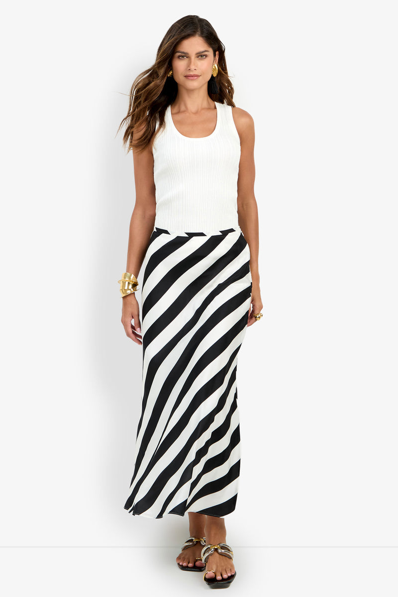 A woman stands against a plain white background wearing a white sleeveless top paired with the Rabina Skirt, a long black-and-white diagonal striped skirt, accessorized with gold bracelets and sandals.