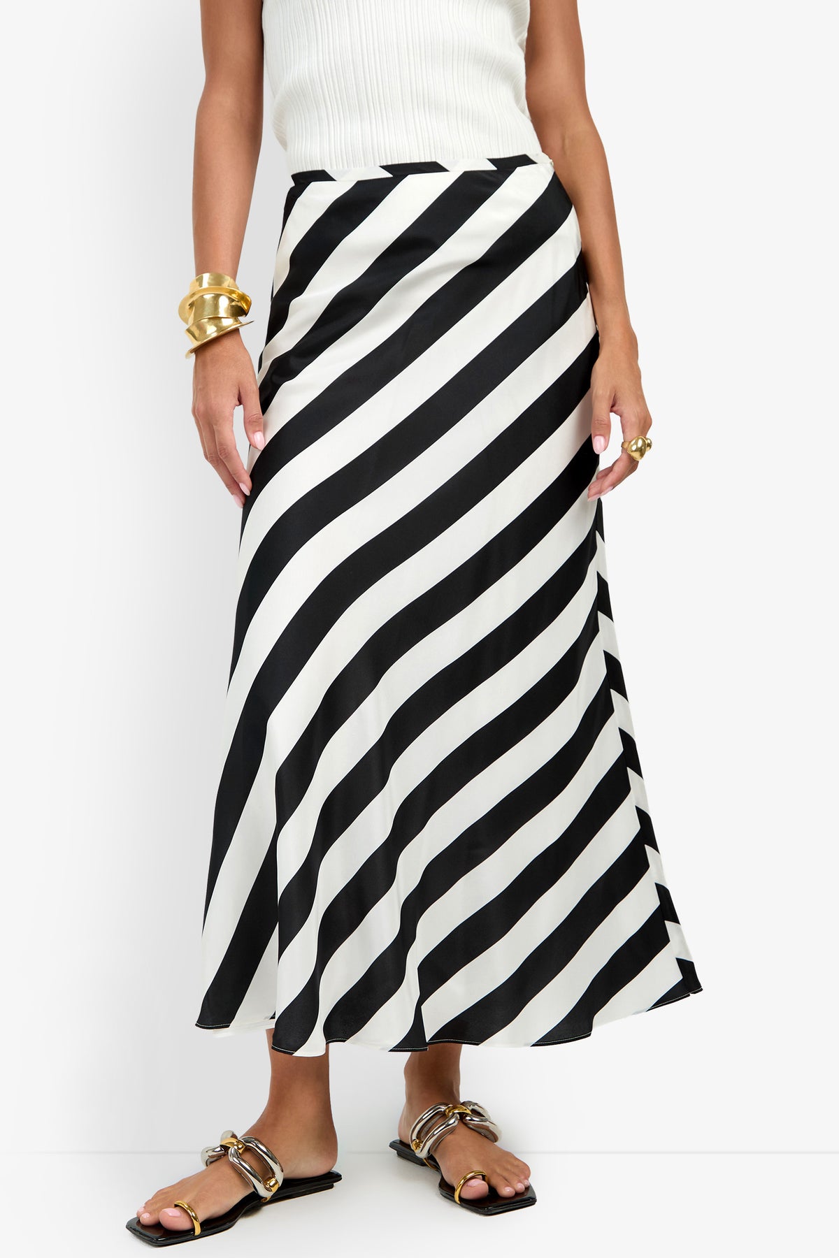 A person wearing a white sleeveless top and the Rabina Skirt—a long black-and-white diagonal striped skirt—accessorized with gold bracelets, rings, and black sandals with gold detail.