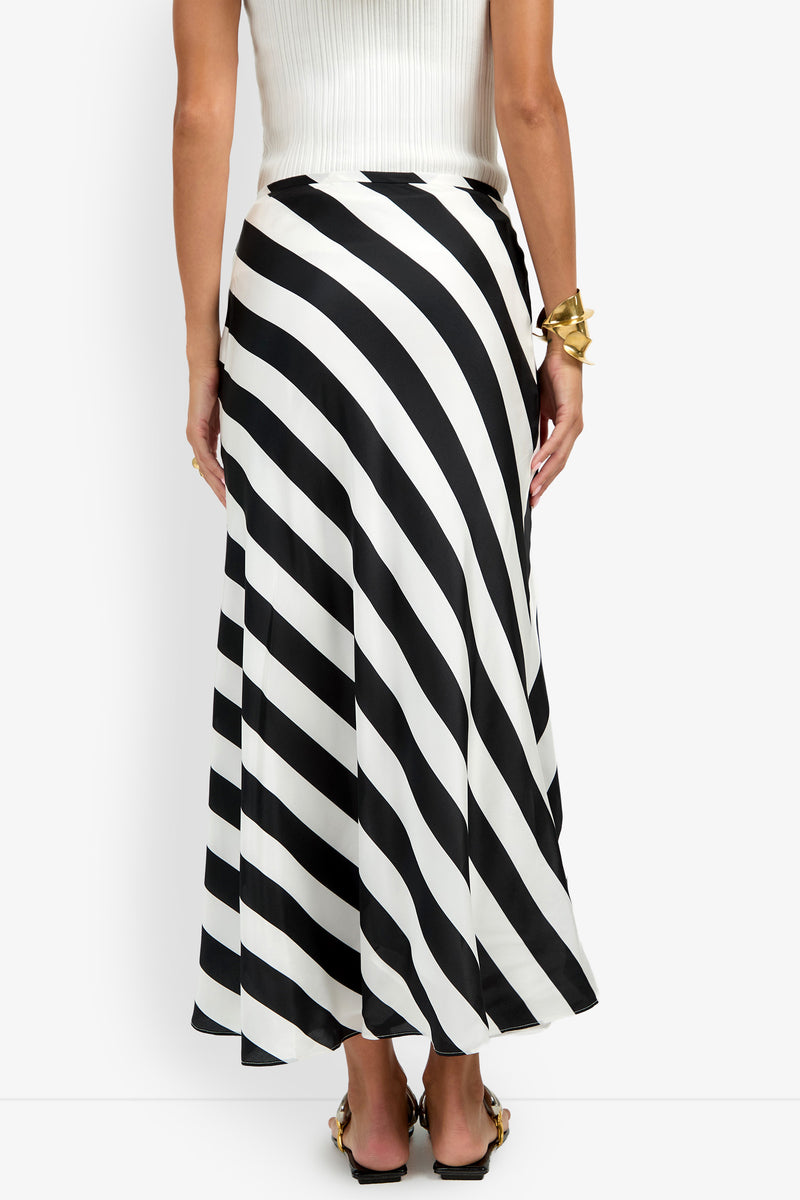 A woman wearing the Rabina Skirt in black and white diagonal stripes, paired with a white ribbed sleeveless top, gold bracelet, and heeled sandals, shown from the back against a plain white background.