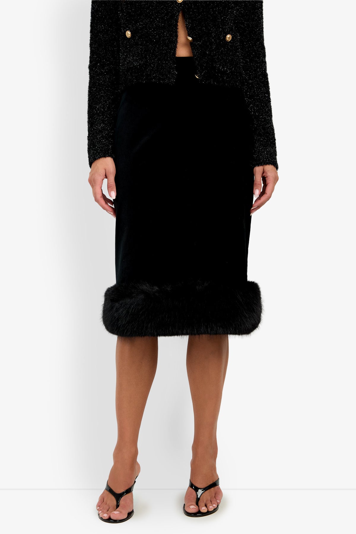 Woman wearing a textured black jacket and the Klara Skirt—a black skirt with a fur trim at the hem—paired with black heeled sandals, standing against a white background.