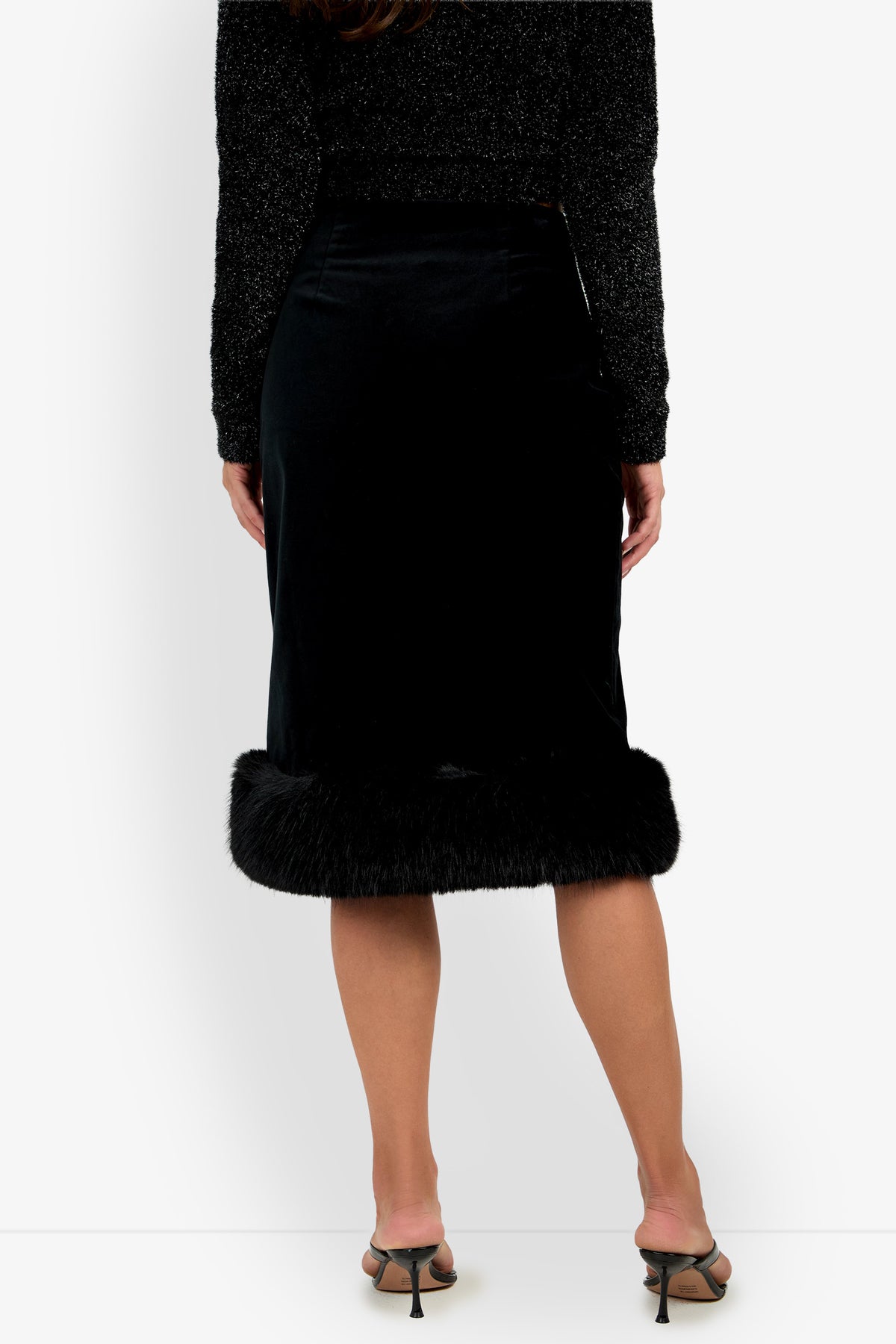 A woman stands facing away, wearing the Klara Skirt—a black midi skirt with a fur trim at the hem—paired with a sparkly black long-sleeve top and open-toe heels. Only her body from shoulders to feet is visible.