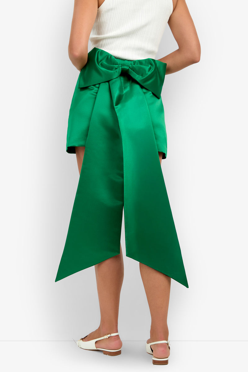 A person wearing a white sleeveless top and the Vinna Skirt—a green high-waisted skirt featuring a large, dramatic satin bow with long tails at the back—paired with white sandals.