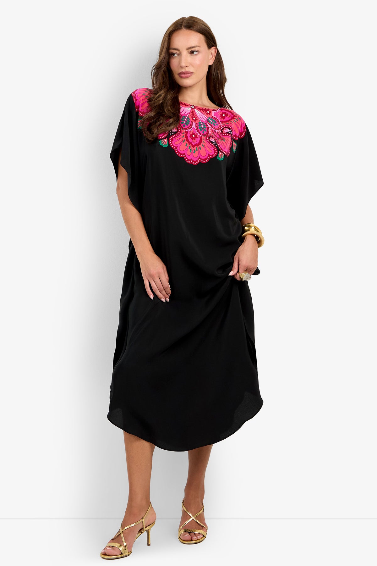 A woman stands against a plain white background wearing the Jalani Maxi Caftan Dress, a loose black design featuring bright pink and red floral embroidery at the neckline. She accessorizes with gold earrings, a gold bracelet, and gold strappy heels.