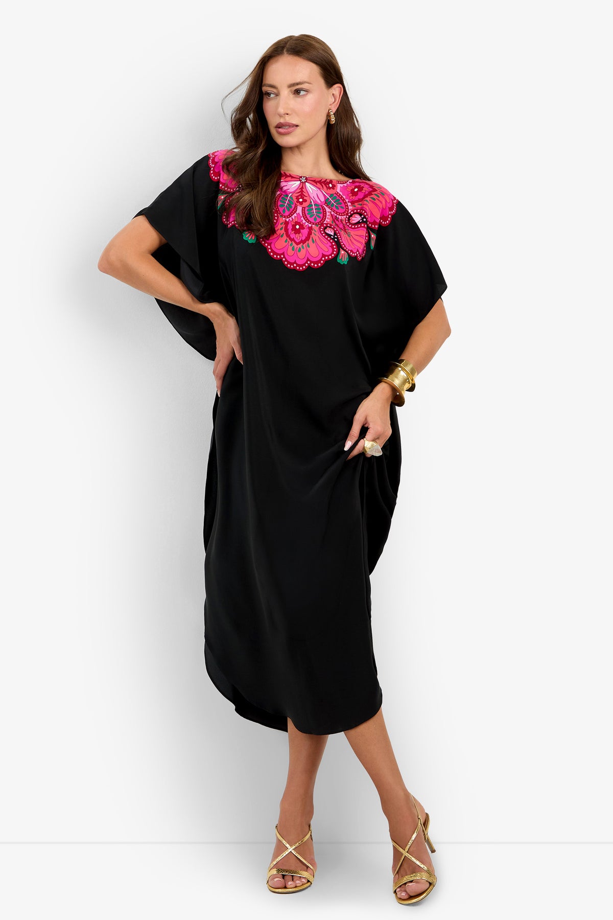 A woman wearing the Jalani Maxi Caftan Dress in Sautoir—a loose black dress featuring a vibrant pink and purple embroidered floral design at the neckline—paired with gold hoop earrings, gold bangles, and beige strappy heels, stands against a plain white background.