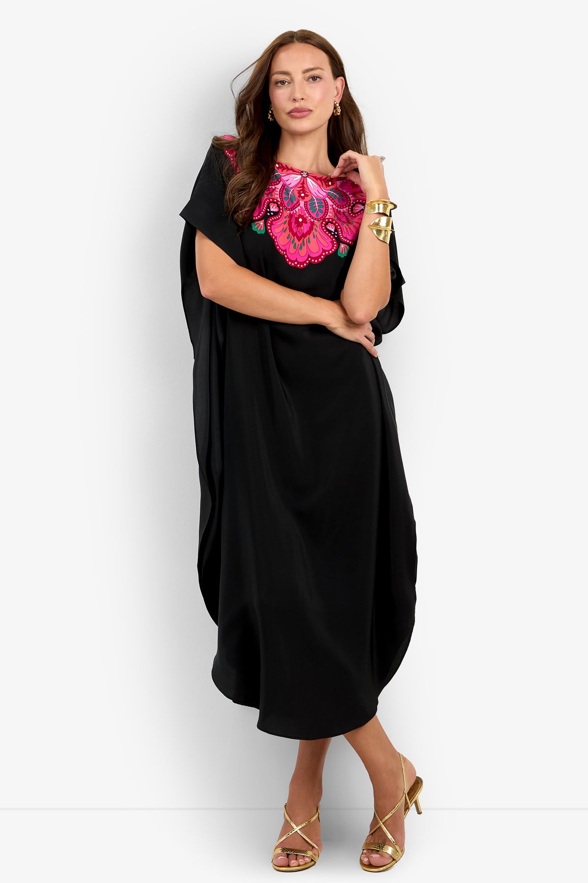 A woman wearing the Jalani Maxi Caftan Dress— a loose black dress featuring a bold pink floral design on the chest— paired with gold earrings, bracelets, and strappy heels, stands with one leg crossed in front of the other against a white background.