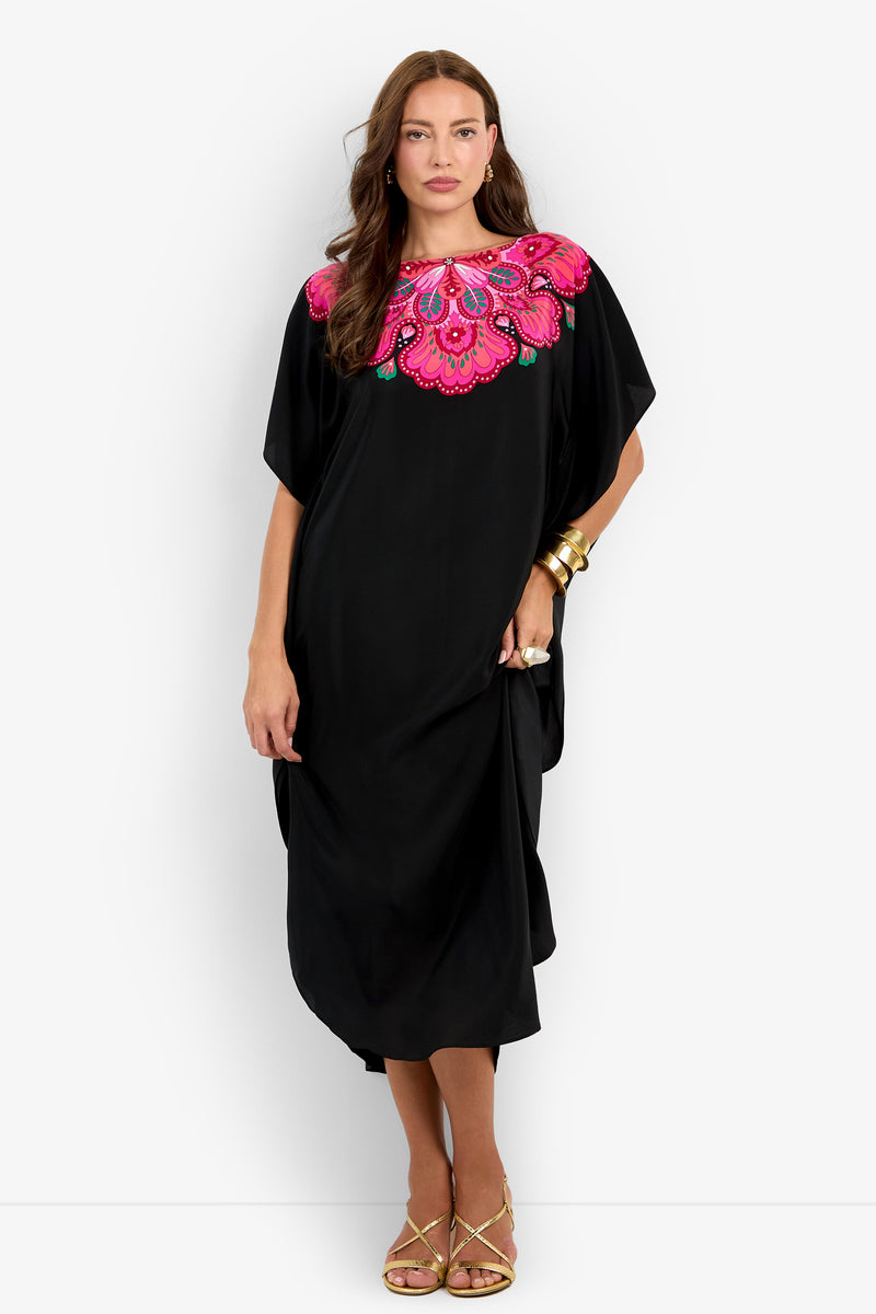 A woman stands with one hand on her hip wearing the Jalani Maxi Caftan Dress, a loose black dress featuring a bright pink embroidered neckline, styled with gold bangles and sandals against a plain white background.