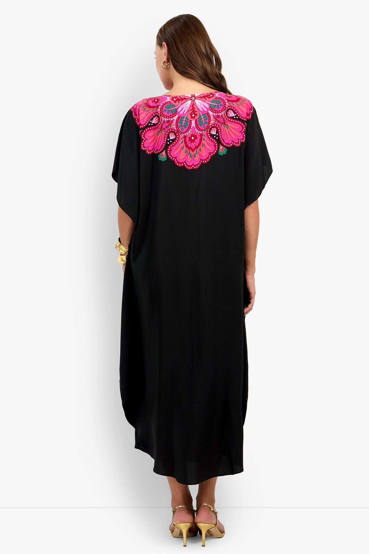 A woman with long brown hair, wearing yellow heels, is seen from behind in the Jalani Maxi Caftan Dress—a long black dress featuring vibrant pink, purple, and green embroidered floral designs on the upper back.