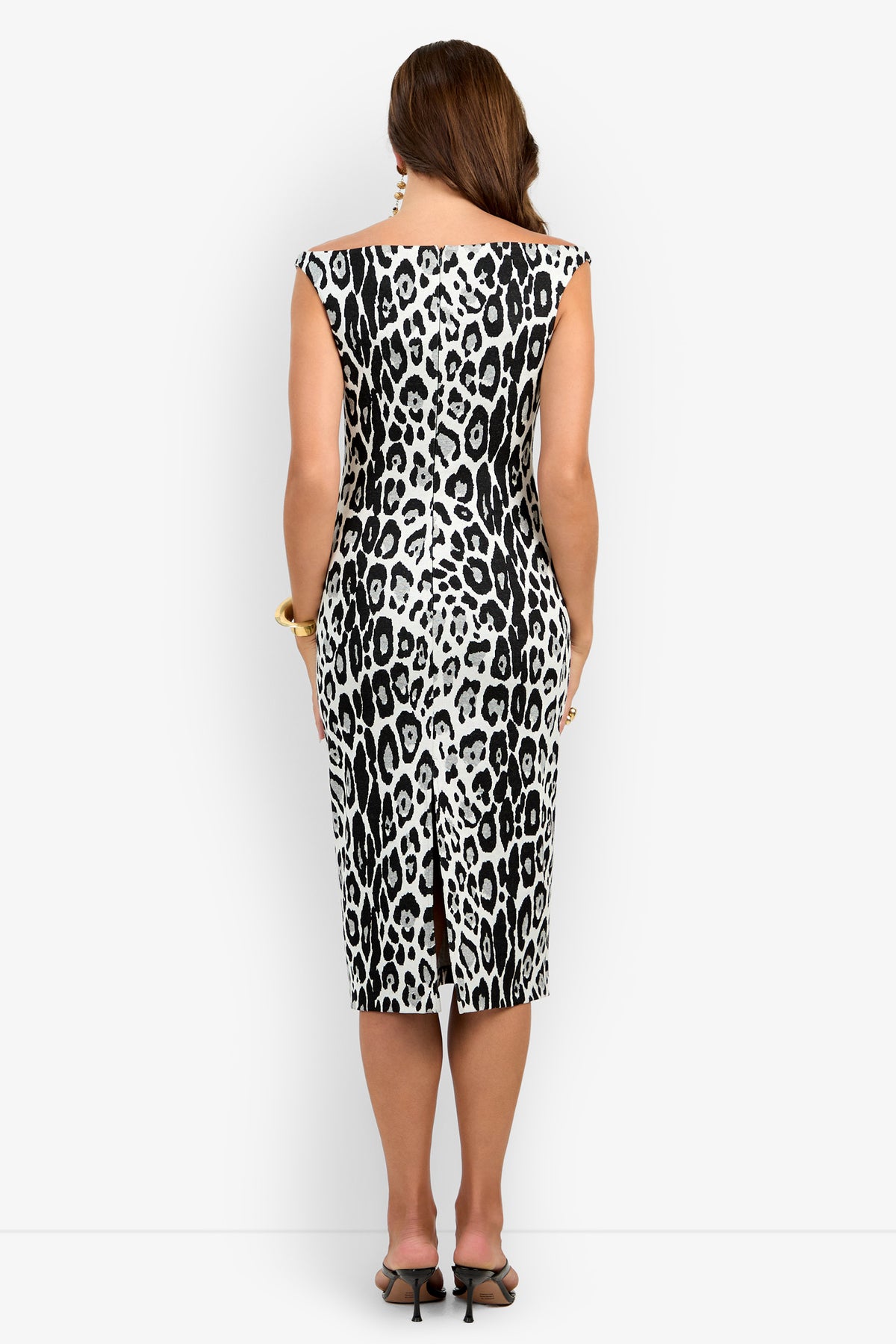 A woman stands facing away, wearing the Emelda Dress—a sleeveless, black and white leopard print, knee-length dress with a back slit. She has long brown hair and pairs the look with black heels and gold accessories.