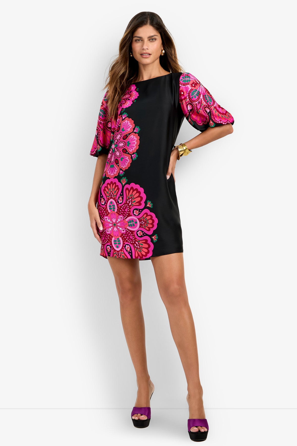 A woman models the Luv Dress—a black mini dress featuring bold pink floral patterns and puffed short sleeves—against a white background, confidently posing with one hand on her hip.
