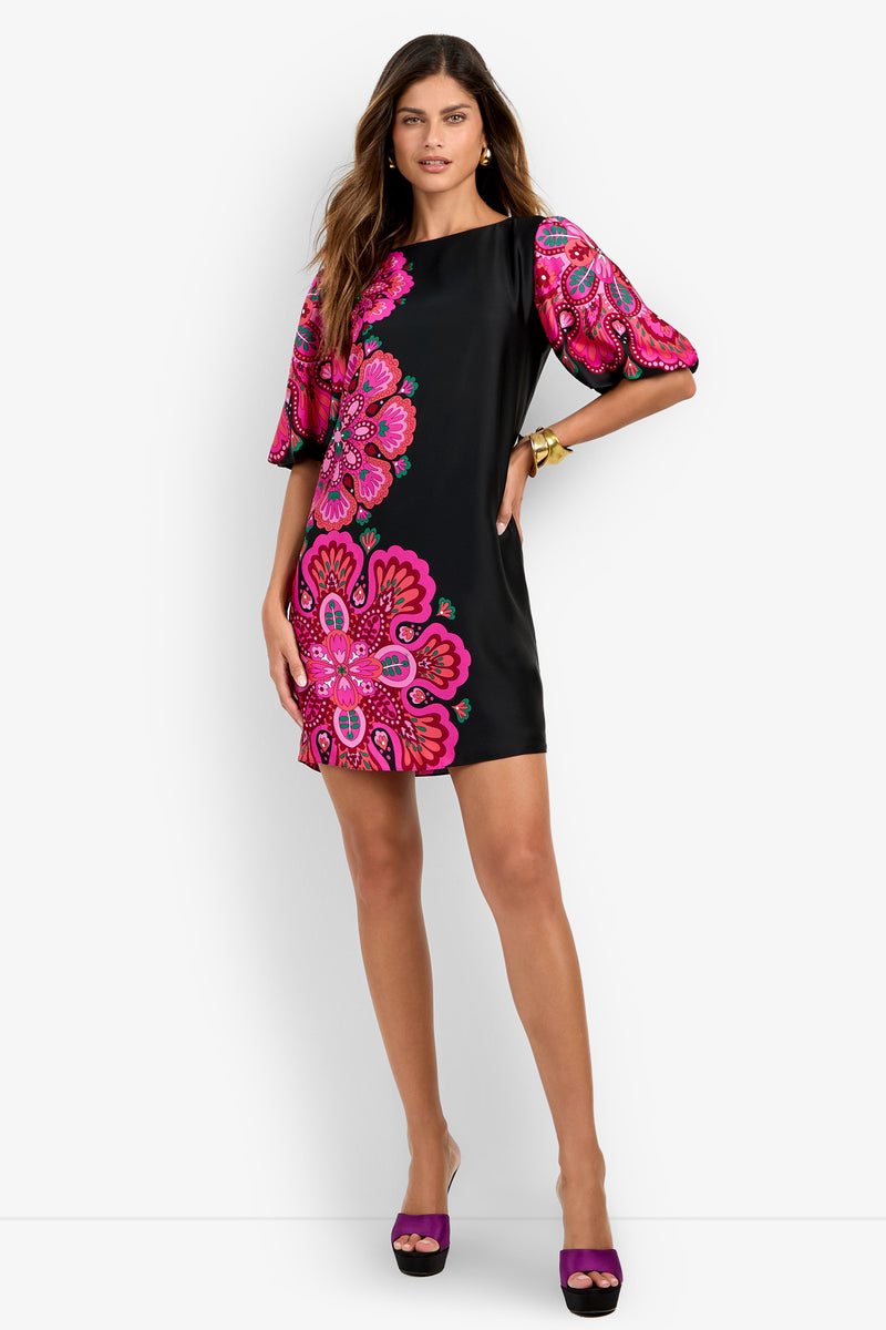 A woman models the Luv Dress—a black mini dress featuring bold pink floral patterns and puffed short sleeves—against a white background, confidently posing with one hand on her hip.