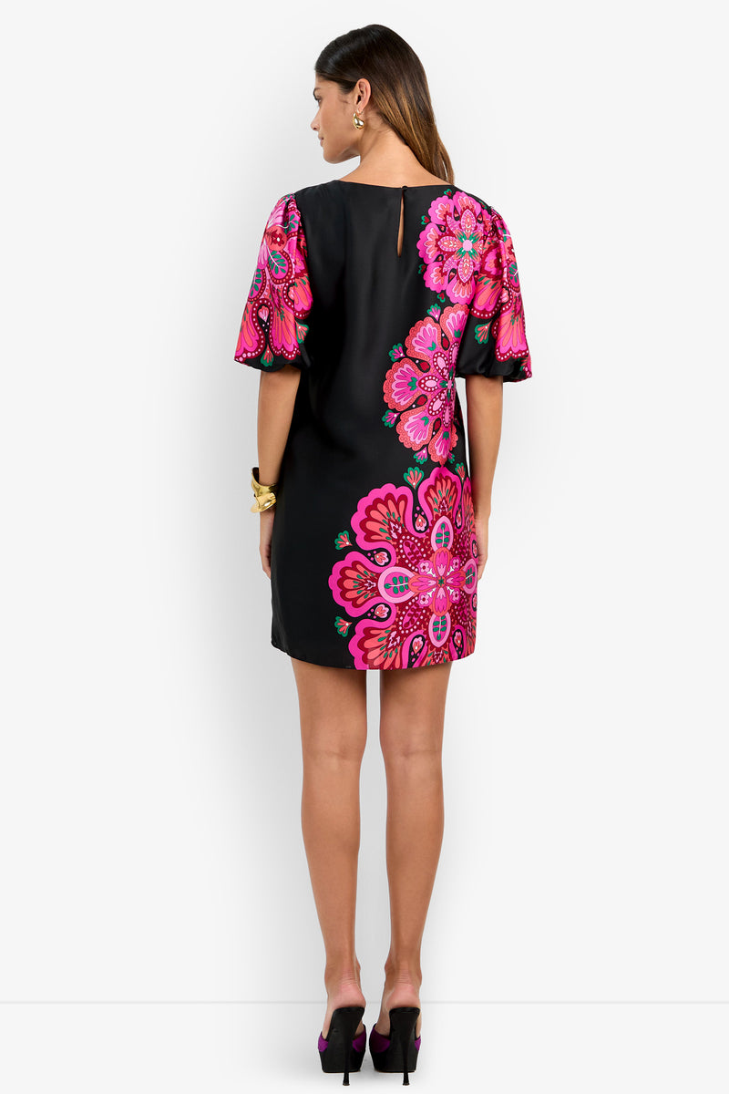 A woman stands with her back to the camera, wearing the Luv Dress— a black dress featuring vibrant pink floral patterns and short sleeves—paired with black heels. She accessorizes with earrings and a bracelet.