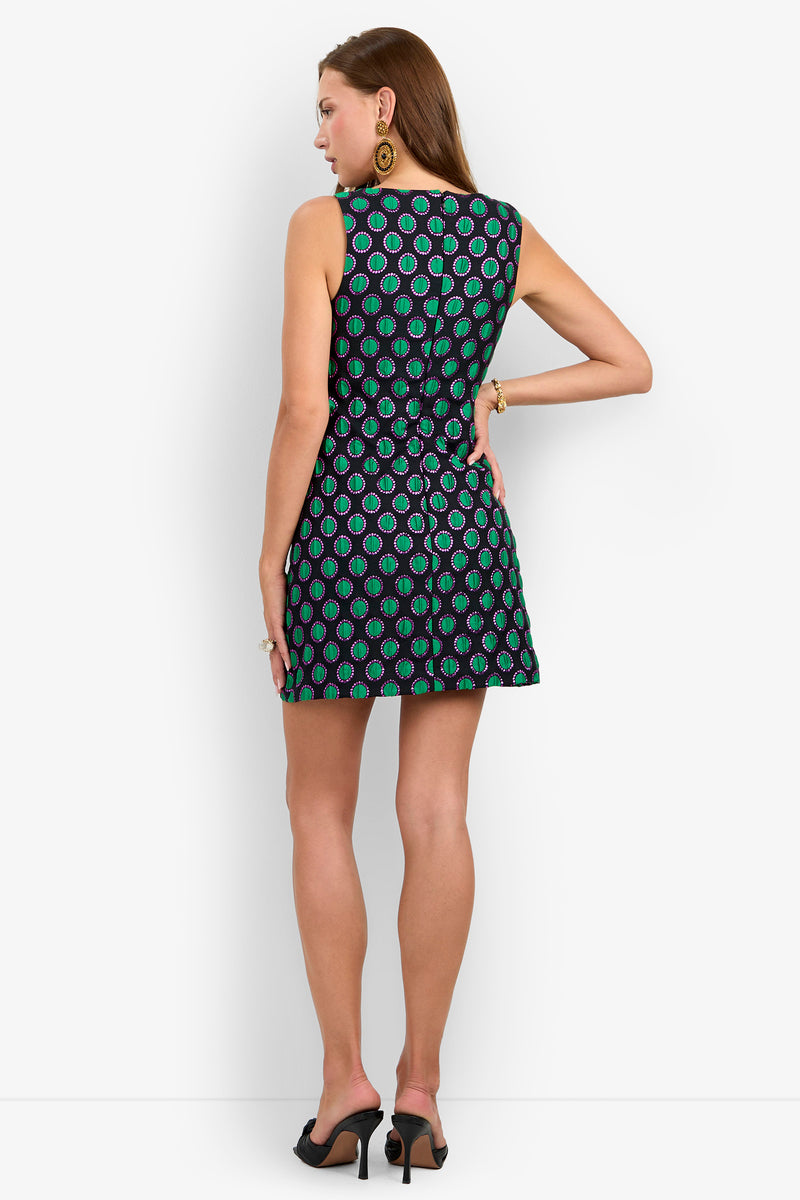 A woman stands with her back to the camera, wearing the BAMBILLA DRESS—a sleeveless, short black dress featuring a green and blue circular pattern woven with subtle metallic fibers—paired with black heels and gold earrings, set against a plain white background.