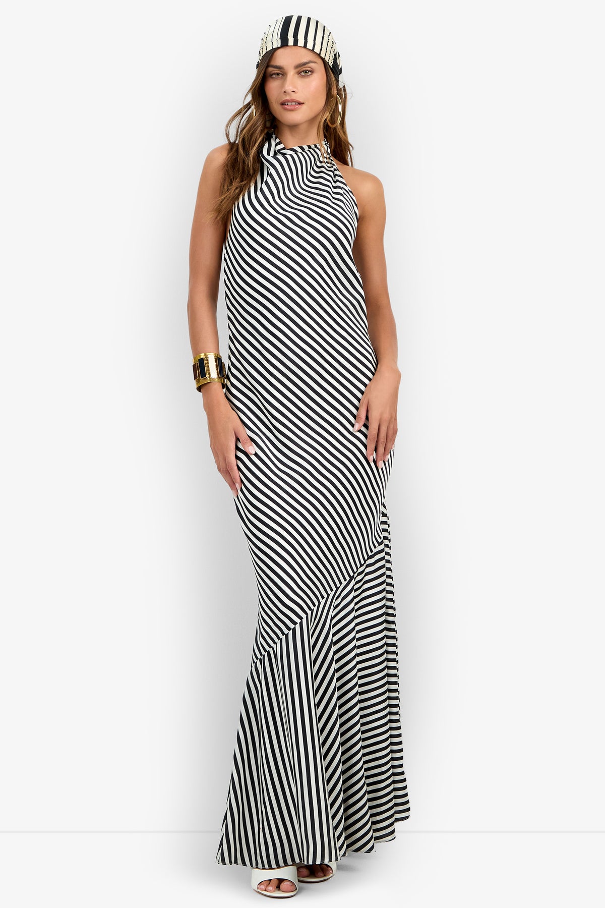 A woman is wearing the Lady Dress—a black and white diagonal striped halter maxi dress with a matching headscarf—paired with white sandals and a gold cuff bracelet, standing against a plain white background.