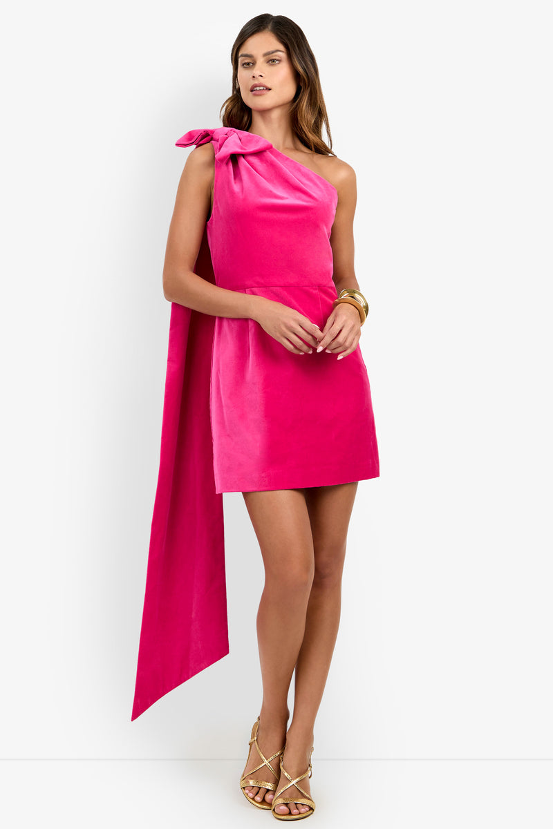 A woman wearing the Romance Dress—a bright pink, one-shoulder mini dress with a large bow and long sash—paired with gold strappy sandals and gold bangles, stands against a white background.