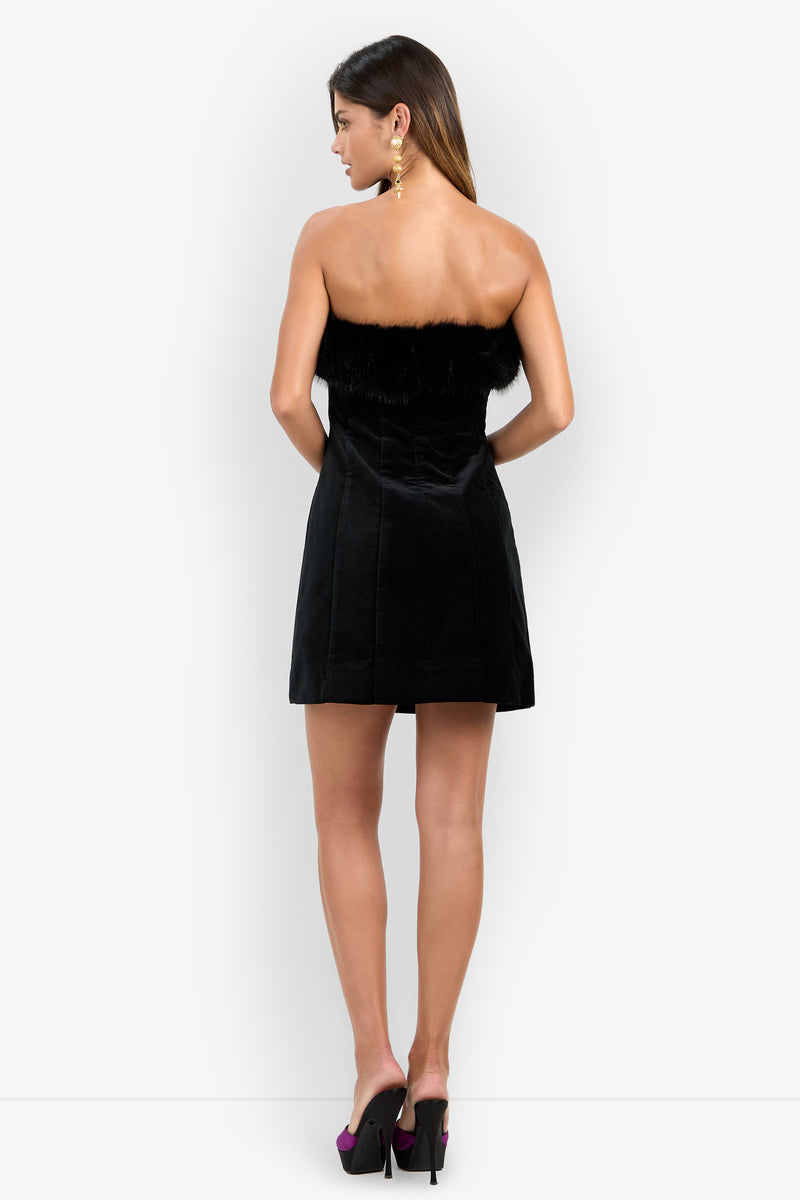 A woman with straight hair and statement earrings stands against a white background, shown from the back, wearing the strapless black Finale Fur Dress with a fur trim and black high heels.