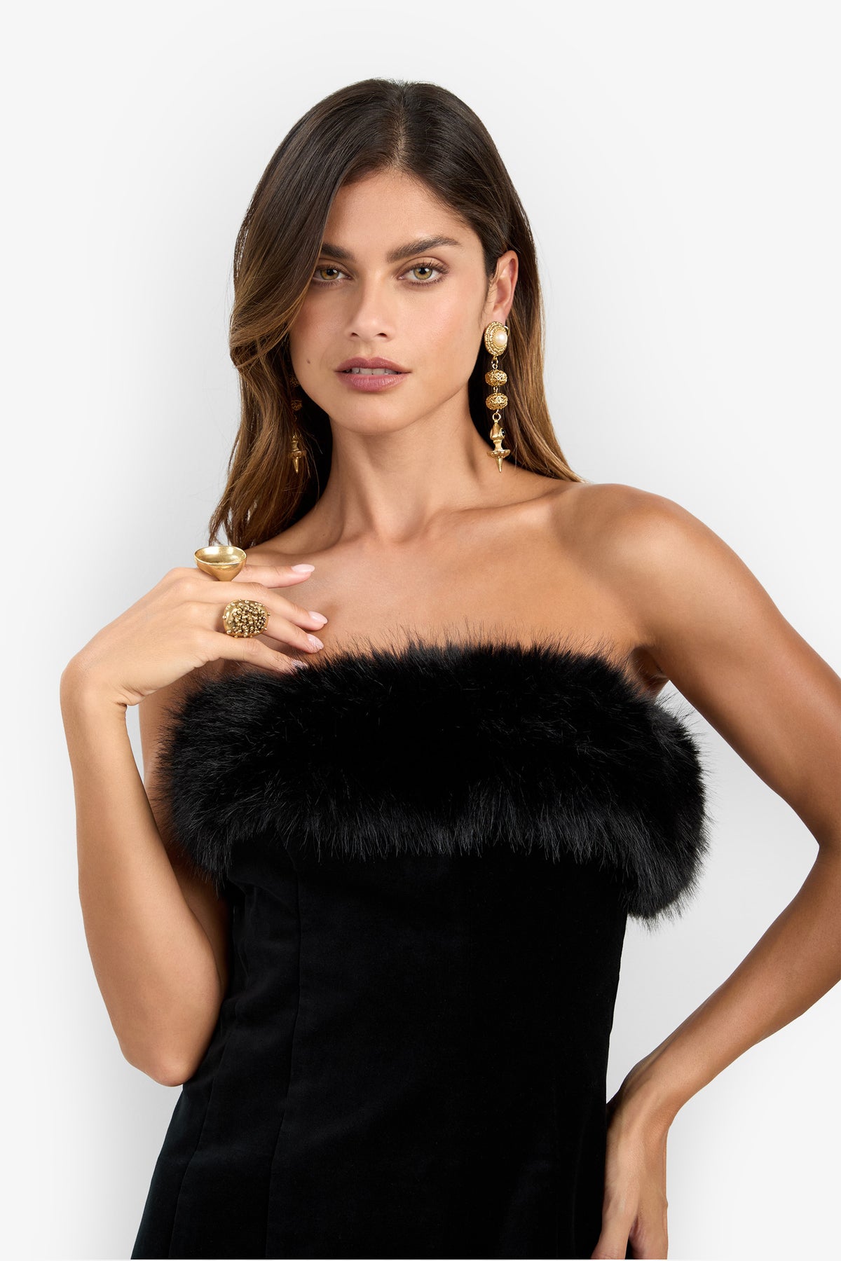 A woman wearing the Finale Fur Dress—a strapless black design with a fur trim—stands against a plain white background, accessorized with large gold earrings and a statement ring as she poses with one hand near her chest.