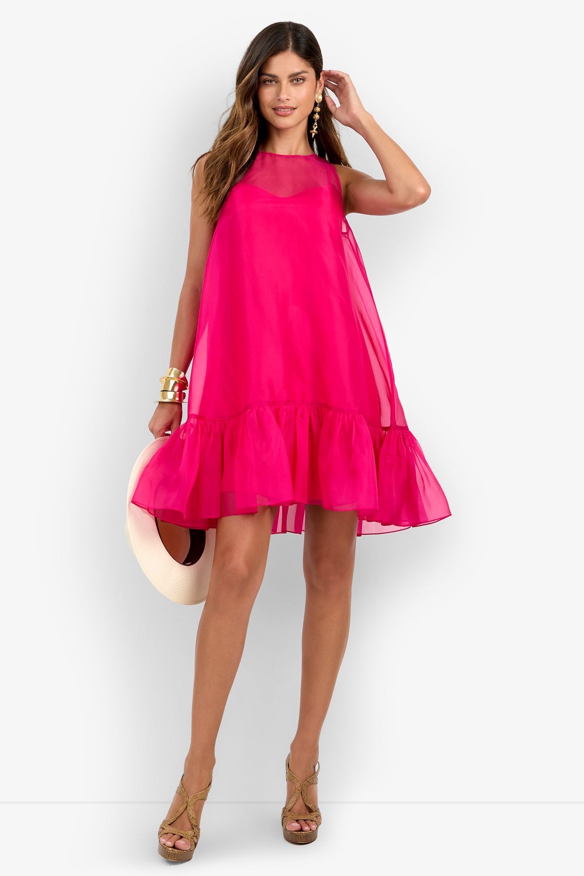 A woman wearing the Winner Dress—a bright pink sleeveless dress with a ruffled hem—paired with gold high-heeled sandals and statement earrings, holding a wide-brimmed hat by her side and smiling.