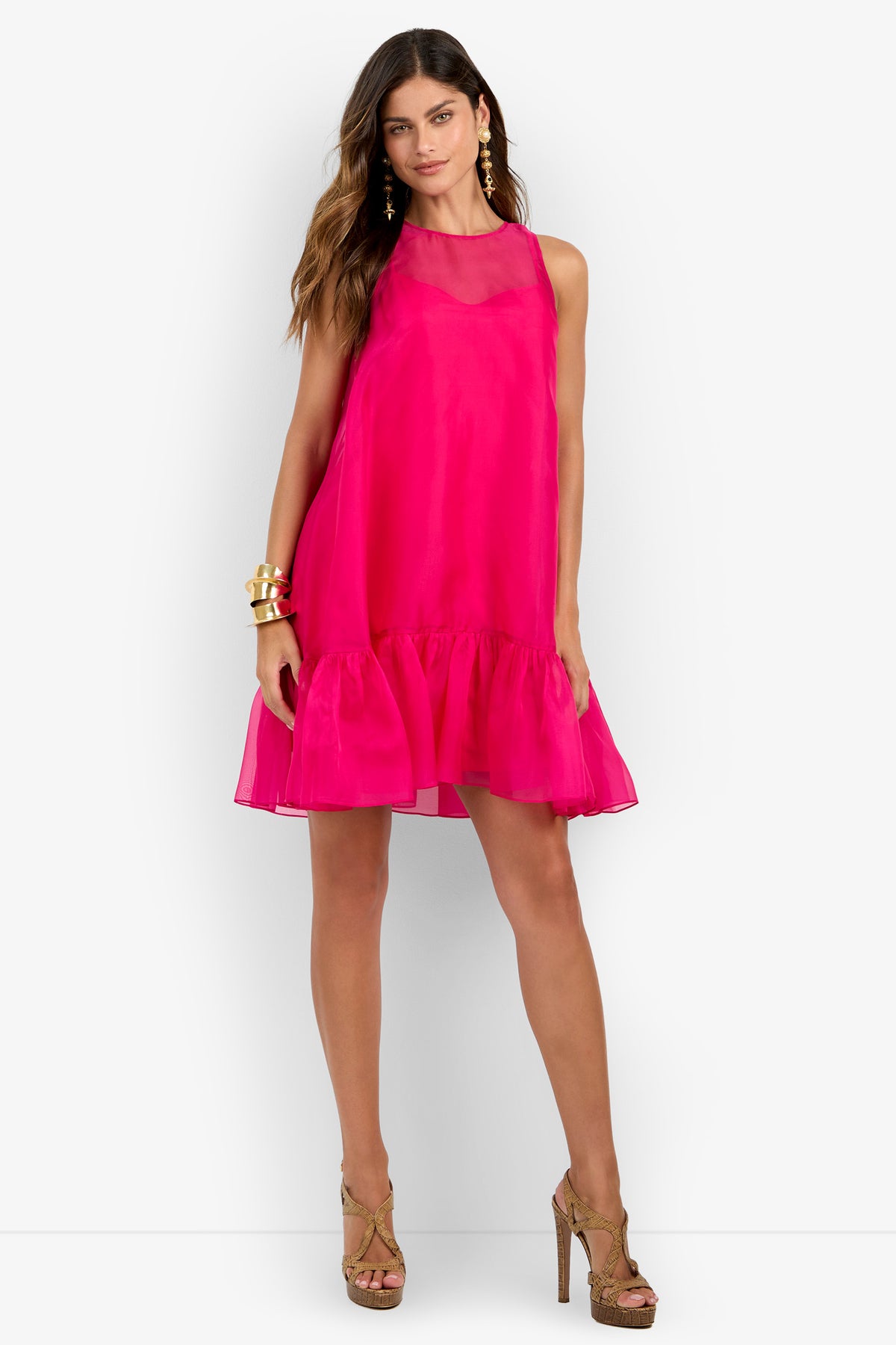A woman wearing the Winner Dress—a bright pink sleeveless ruffled dress—paired with gold high-heeled sandals, gold earrings, and bracelets, stands against a white background.