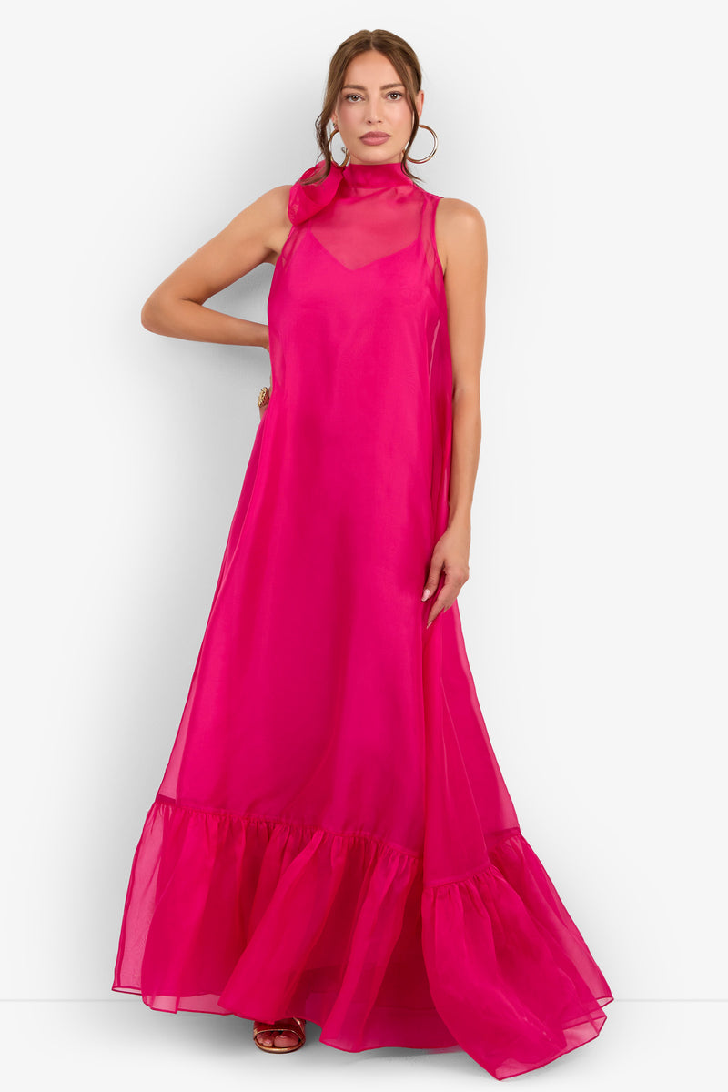 A woman wearing the Leading Dress—a sleeveless, floor-length gown in bright pink featuring a sheer high neckline and ruffled hem—poses with one hand on her hip against a white background.